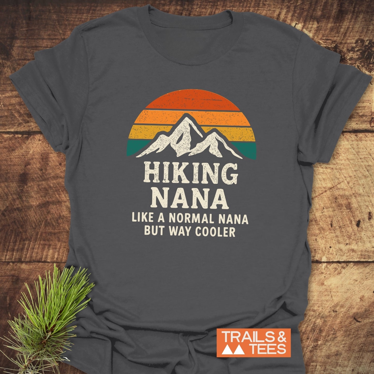 Hiking Nana Funny T-Shirt featuring a mountain silhouette with retro sunset stripes, laid on wood. Includes text Hiking Nana Like a normal Nana but way cooler and Trails & Tees logo.