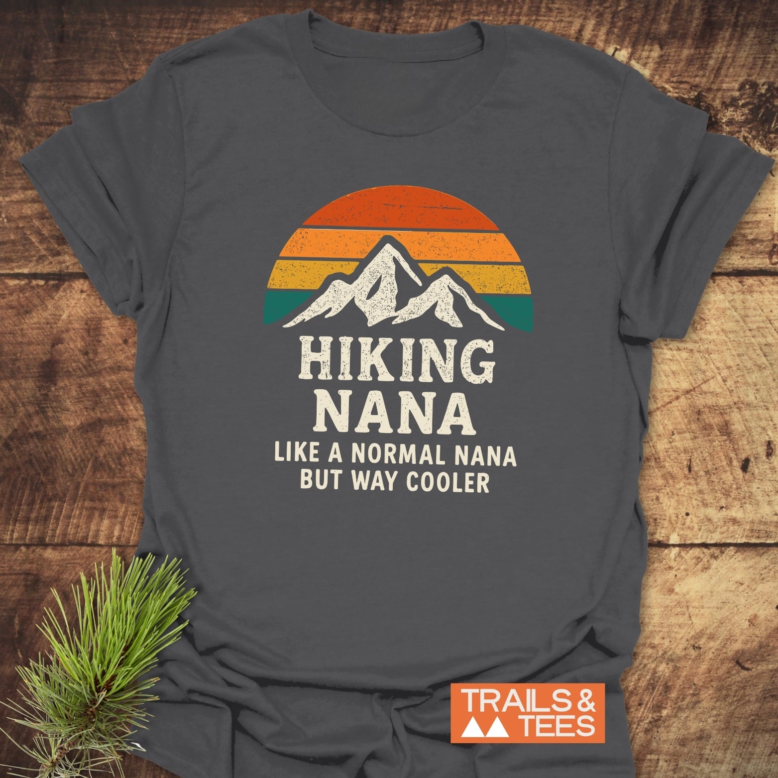 Hiking Nana Funny T-Shirt featuring a mountain silhouette with retro sunset stripes, laid on wood. Includes text Hiking Nana Like a normal Nana but way cooler and Trails & Tees logo.