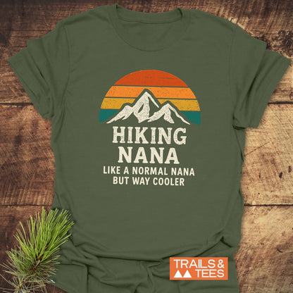 Hiking Nana Funny T-Shirt on wooden surface, featuring a retro sun and mountain graphic. Text reads: HIKING NANA Like a normal Nana but way cooler. Includes Trails & Tees logo.