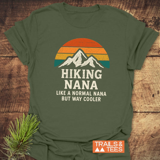 Hiking Nana Funny T-Shirt on wooden surface, featuring a retro sun and mountain graphic. Text reads: HIKING NANA Like a normal Nana but way cooler. Includes Trails & Tees logo.
