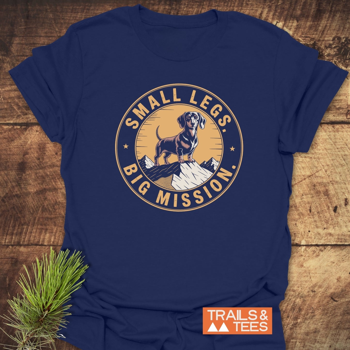 Hiking Dachshound/Wiener Dog T-Shirt features a vintage-style graphic of a cartoon dachshund on a snowy peak with the slogan “SMALL LEGS, BIG MISSION.” Includes “TRAILS & TEES” logo.