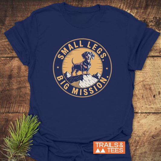 Hiking Dachshound/Wiener Dog T-Shirt features a vintage-style graphic of a cartoon dachshund on a snowy peak with the slogan “SMALL LEGS, BIG MISSION.” Includes “TRAILS & TEES” logo.