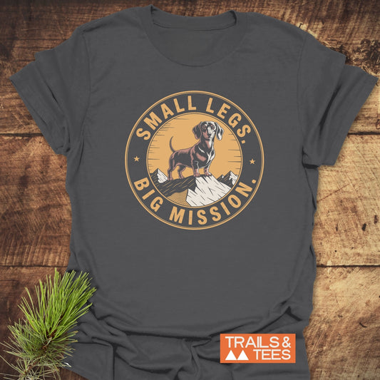 Charcoal-gray Hiking Dachshound/Wiener Dog T-Shirt with dachshund and mountain badge graphic, SMALL LEGS. BIG MISSION. text, and TRAILS & TEES logo, laid on wooden surface.