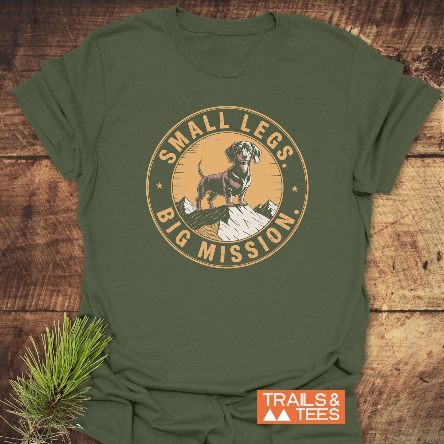 Hiking Dachshound/Wiener Dog T-Shirt featuring a vintage-style dachshund graphic on a snowy peak with SMALL LEGS, BIG MISSION text, and TRAILS & TEES logo on olive-green fabric.