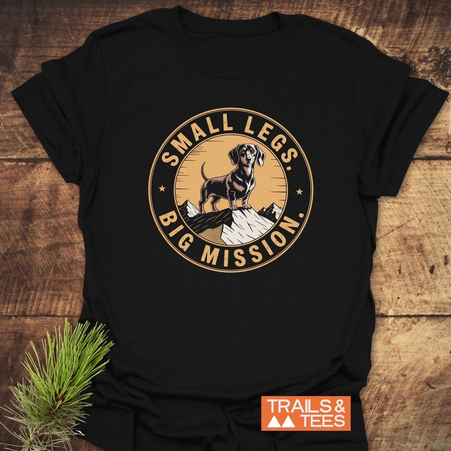 Hiking Dachshund/Wiener Dog T-Shirt featuring a dachshund graphic atop a mountain with “SMALL LEGS, BIG MISSION.” Laid on wood, includes “TRAILS & TEES” logo, emphasizing outdoor adventure spirit.