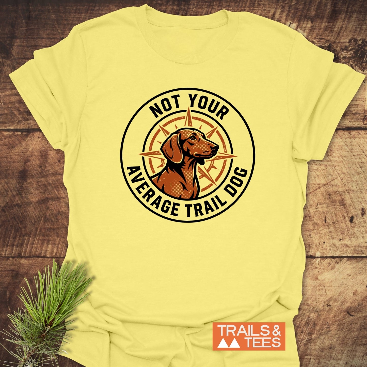 Not Your Average Trail Dog Wiener T-Shirt featuring a dog and compass logo on pale-yellow fabric, laid flat on wood. Includes TRAILS & TEES label, embodying outdoor adventure and comfort.