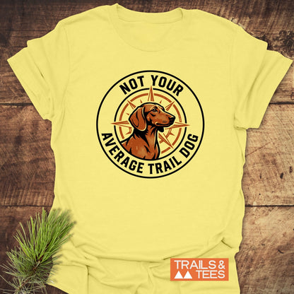 Not Your Average Trail Dog Wiener T-Shirt featuring a dog and compass logo on pale-yellow fabric, laid flat on wood. Includes TRAILS & TEES label, embodying outdoor adventure and comfort.