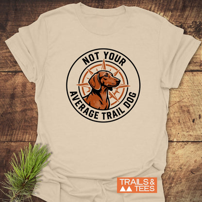 Light-beige Not Your Average Trail Dog Wiener T-Shirt with a black-and-orange logo featuring a trail dog over a compass rose, laid on wood; branded TRAILS & TEES.