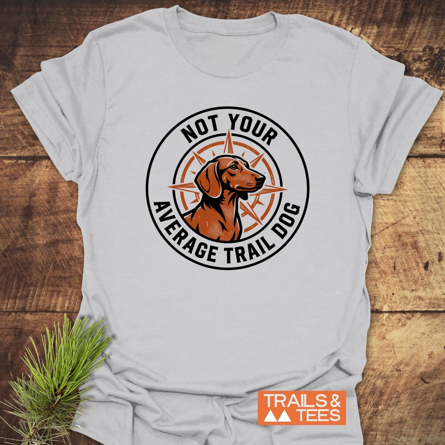 Not Your Average Trail Dog Wiener T-Shirt featuring a brown trail dog logo on a light-gray shirt. It showcases a compass rose design with the phrase encircling the image, embodying outdoor adventure spirit.