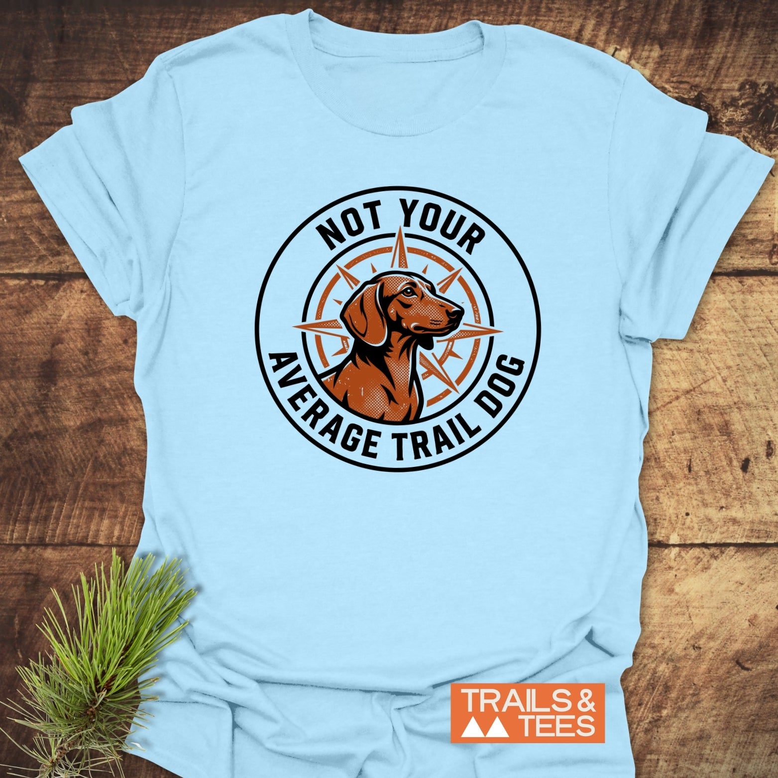 Not Your Average Trail Dog Wiener T-Shirt featuring a trail dog and compass badge design, laid flat on wood with a pine sprig. Includes “TRAILS & TEES” logo near hem.