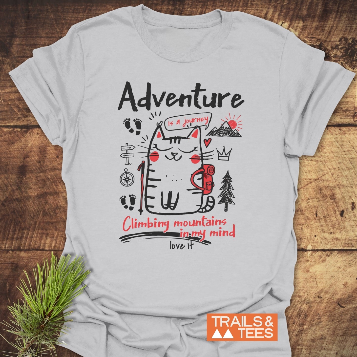 Adventure Cat Funny T-Shirt featuring a cartoon cat with a backpack and trekking pole, surrounded by playful doodles. Adventure is a journey theme, with TRAILS & TEES logo visible.