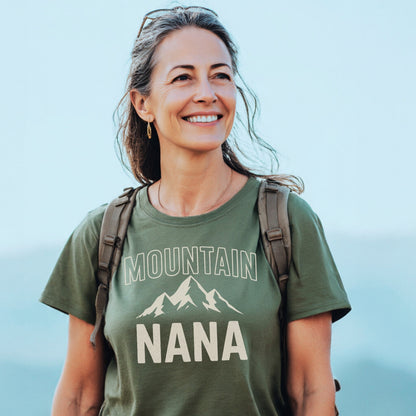 Woman outdoors wearing Mountain Nana T-Shirt with mountain graphic, light backpack, and sunglasses on head, smiling amidst a pale sky, embodying the tee's trail-tested comfort and timeless design.