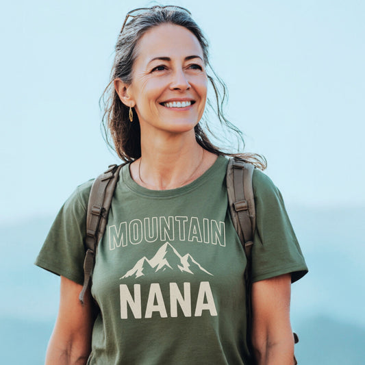 Woman outdoors wearing Mountain Nana T-Shirt with mountain graphic, light backpack, and sunglasses on head, smiling amidst a pale sky, embodying the tee's trail-tested comfort and timeless design.