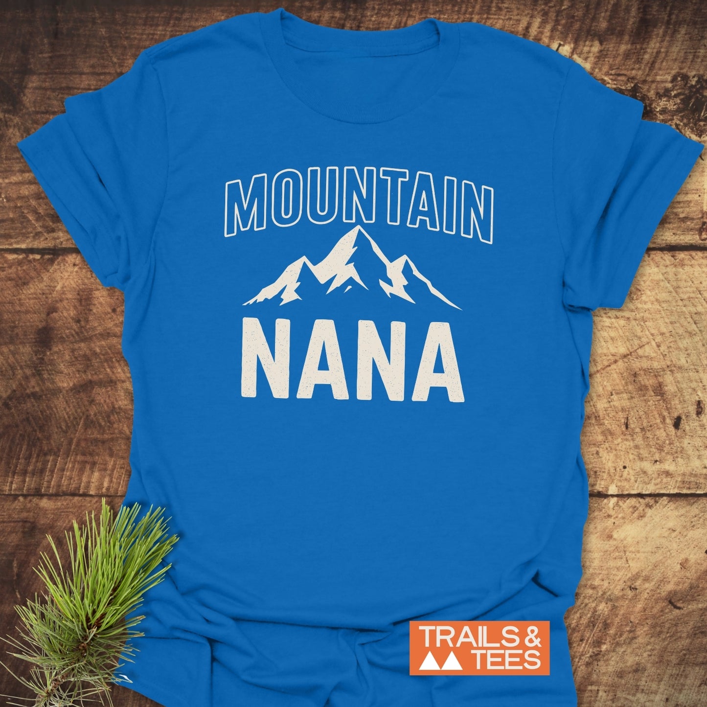Mountain Nana T-Shirt with MOUNTAIN and NANA printed above a mountain graphic on a bright blue tee, showcasing a rustic touch with a pine sprig and Trails & Tees logo.
