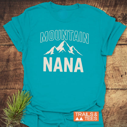Mountain Nana T-Shirt featuring a white mountain graphic, “MOUNTAIN” above, “NANA” below, on a turquoise-blue shirt. Includes TRAILS & TEES logo and pine sprig accent.