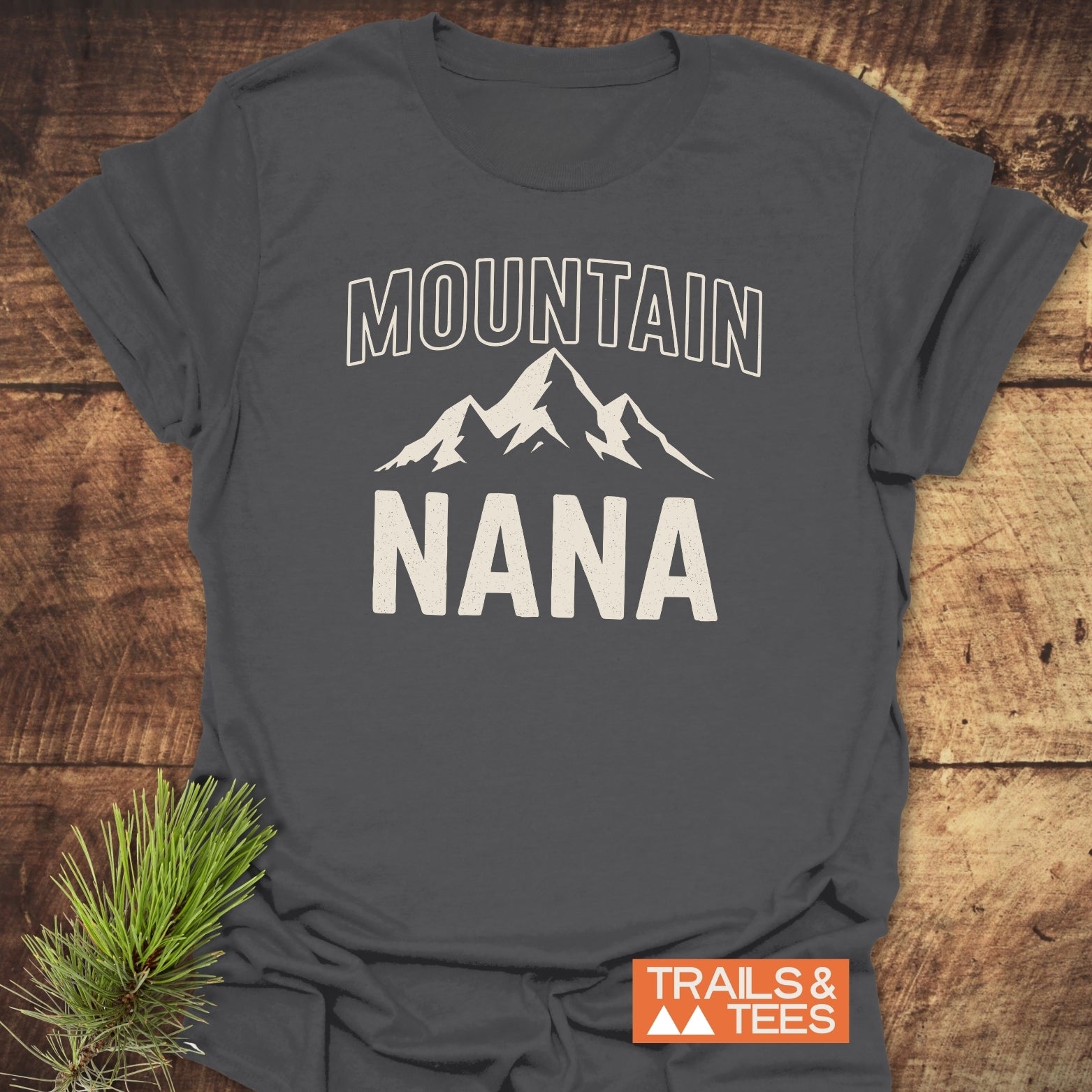 Mountain Nana T-Shirt featuring a stylized mountain graphic, crafted from soft ring-spun cotton. Displayed on a wooden surface with “Trails & Tees” logo and pine branch accent.