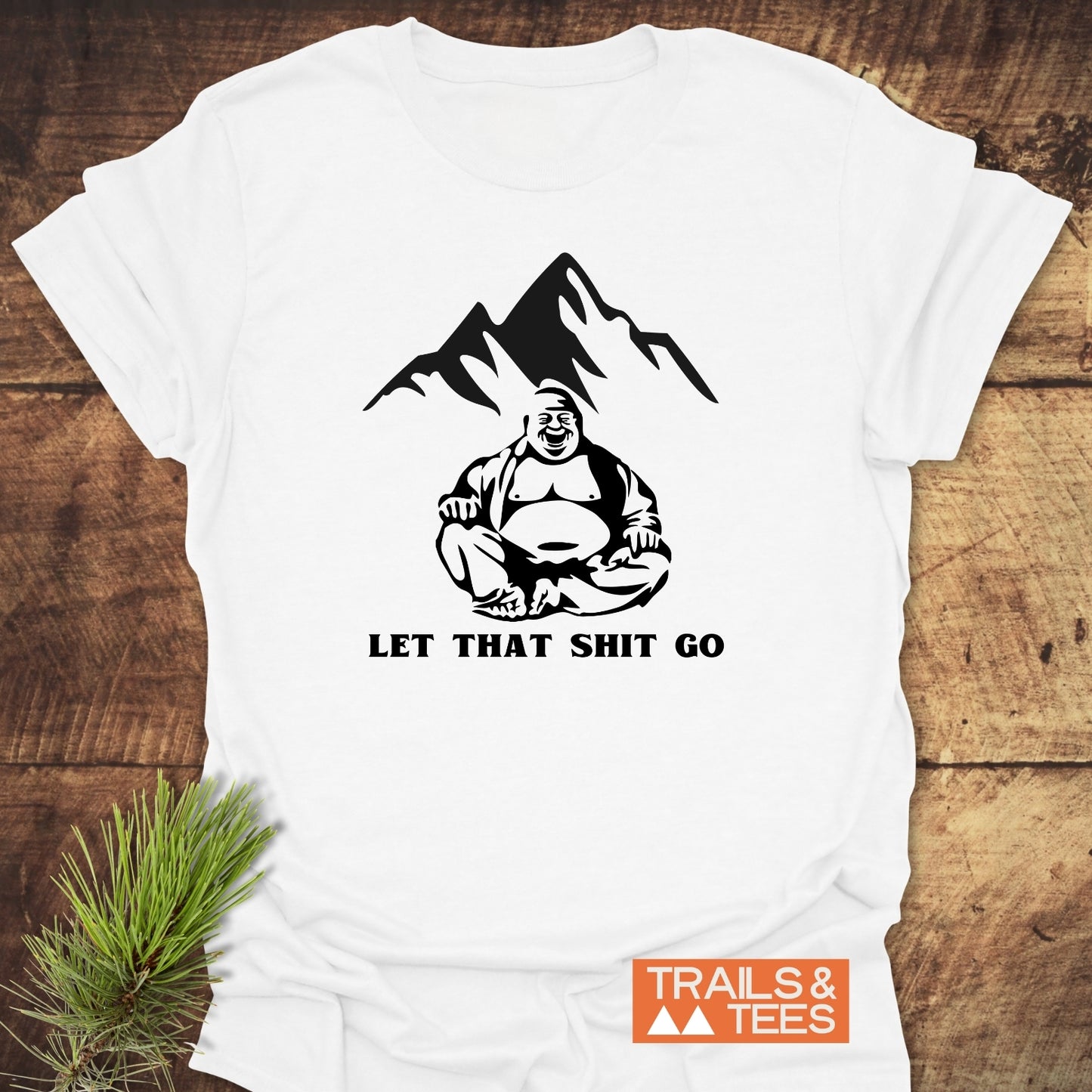 Let That Sh*t Go Funny T-Shirt featuring a laughing Buddha in front of a mountain, laid on wood with pine sprig. Includes “TRAILS & TEES” logo, crafted for outdoor comfort and durability.