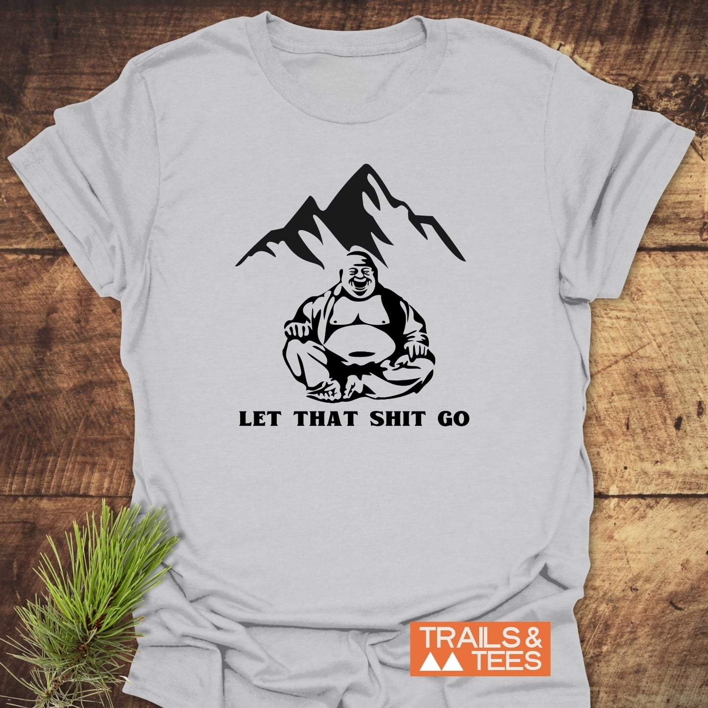 Let That Sh*t Go Funny T-Shirt features a black laughing Buddha graphic with mountains, slogan beneath, on soft cotton. Styled with green sprig, Trails & Tees logo overlay in corner.