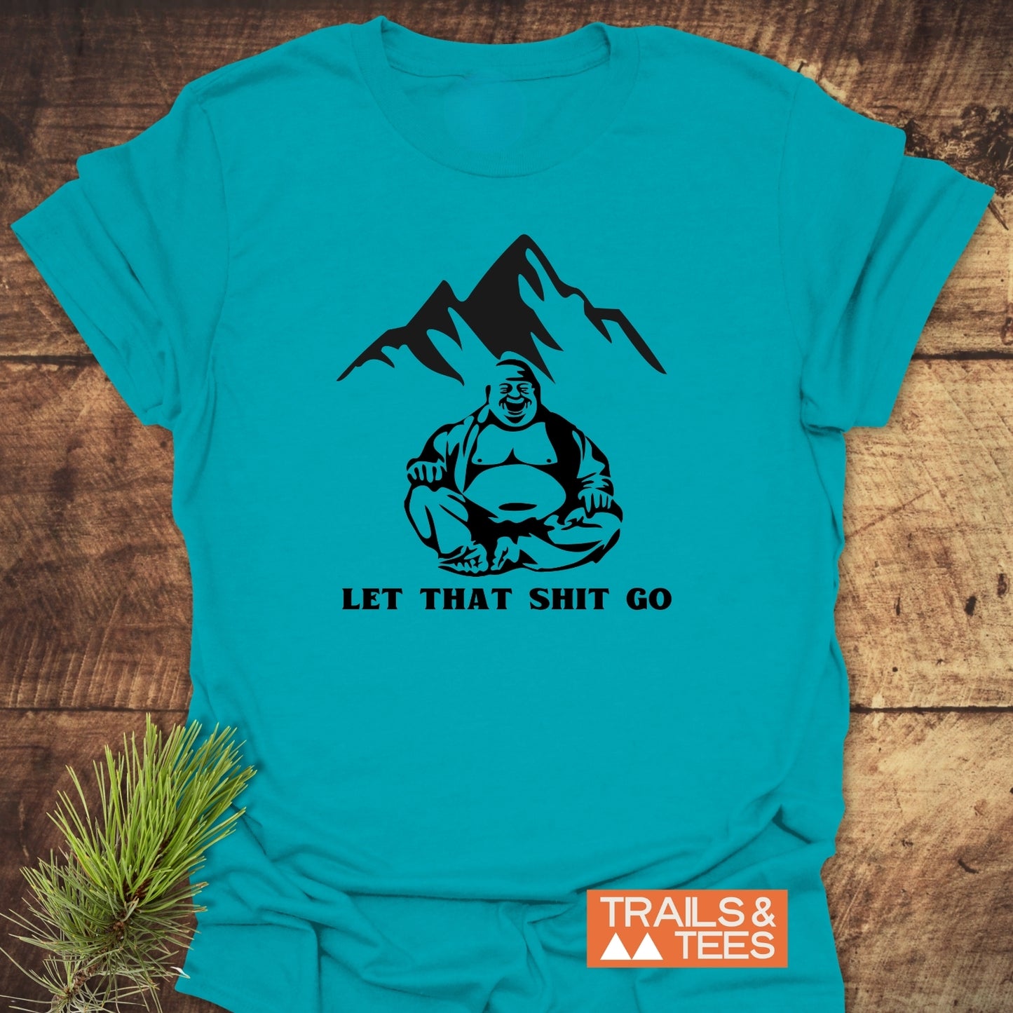 Let That Sh*t Go Funny T-Shirt featuring a mountain and Buddha print on a soft, ring-spun cotton fabric, ideal for outdoor enthusiasts, with a Trails & Tees logo in the corner.