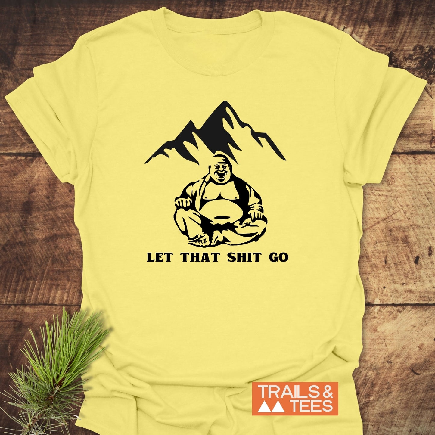 Pale-yellow Let That Sh*t Go Funny T-Shirt featuring a black-line Buddha drawing with mountains and bold text, laid flat on wood, showcasing the TRAILS & TEES logo for an outdoor-inspired vibe.