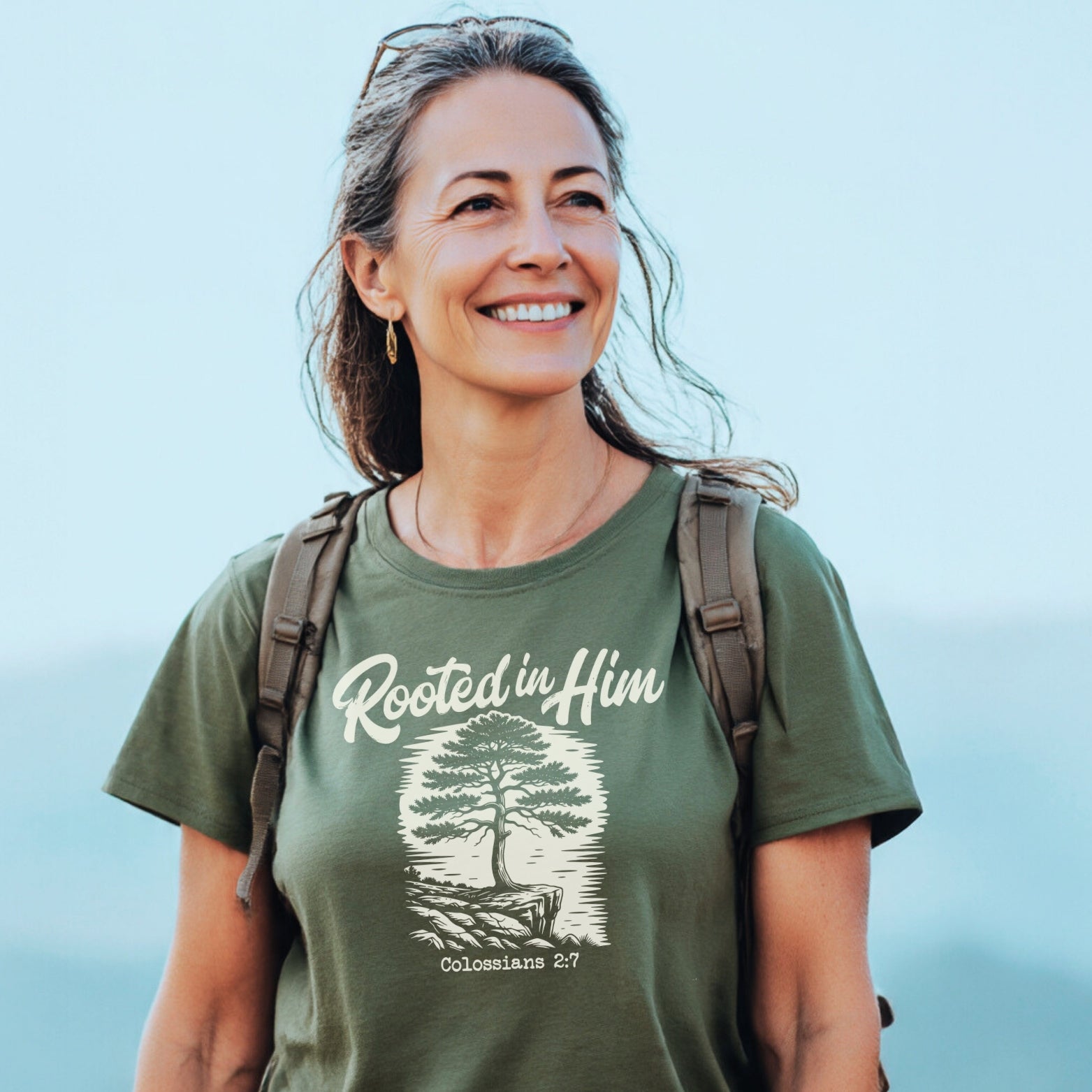 Woman outdoors wearing the Rooted In Him Mountain T-Shirt, featuring a tree illustration with “Colossians 2:7.” She carries a backpack, exuding adventure-ready comfort and style.
