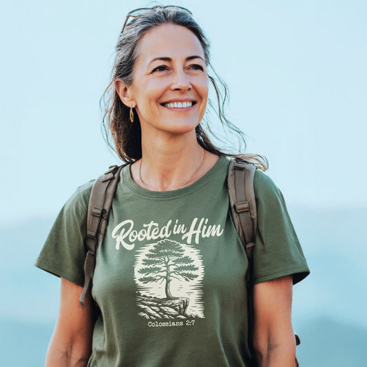 Woman outdoors wearing the Rooted In Him Mountain T-Shirt, featuring a tree illustration with “Colossians 2:7.” She carries a backpack, exuding adventure-ready comfort and style.