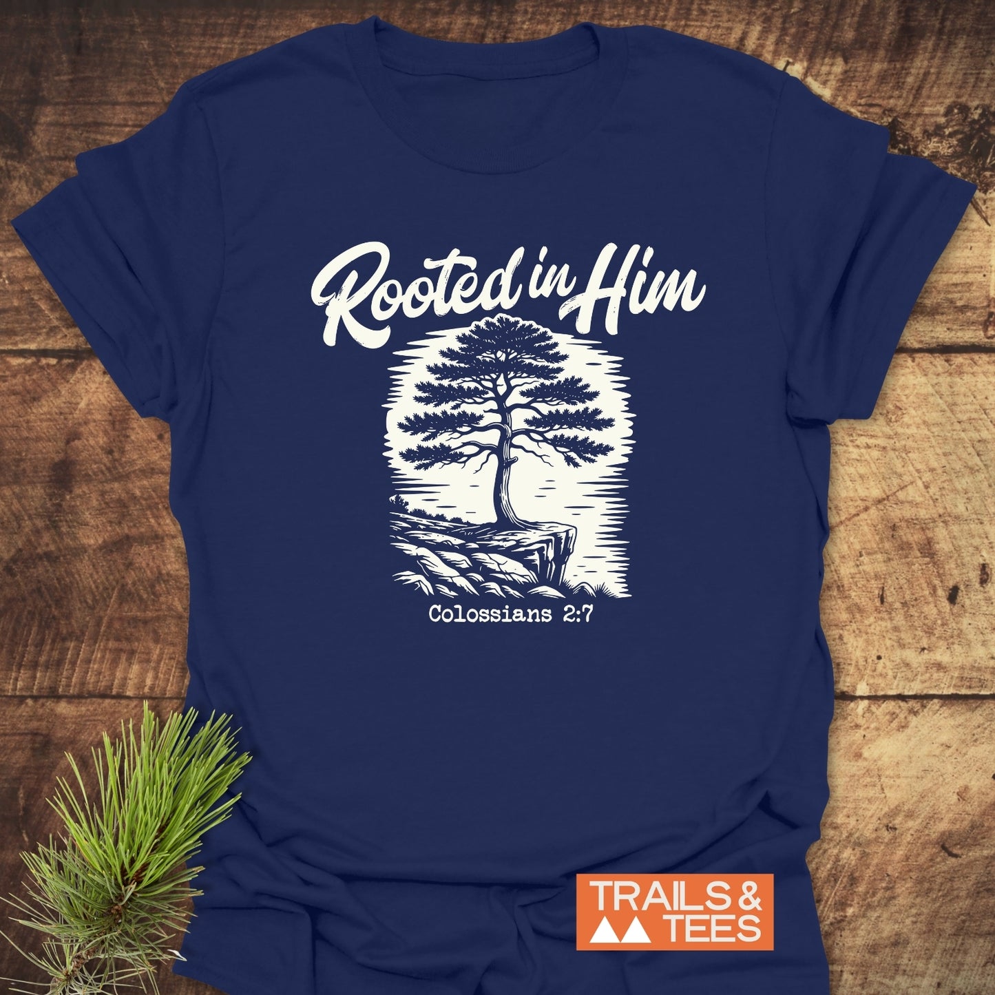 Rooted In Him Mountain T-Shirt features a “Rooted in Him” text and tree graphic with Colossians 2:7, displayed on a navy T-shirt; includes brand logo TRAILS & TEES and pine sprig.