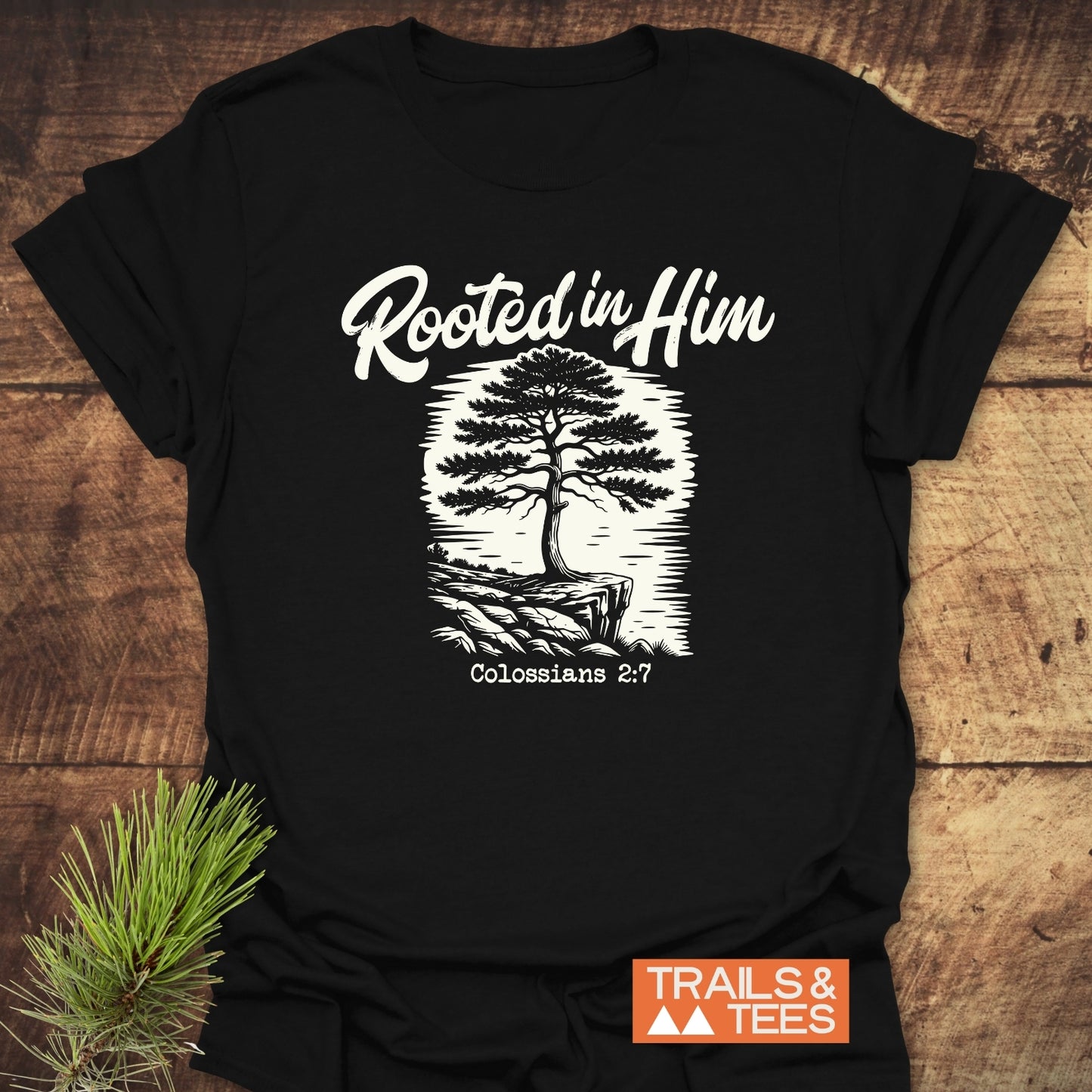 Rooted In Him Mountain T-Shirt featuring a white tree graphic on a cliff with “Rooted in Him” and “Colossians 2:7.” Includes Trails & Tees logo, showcasing trail-tested comfort and durable design.