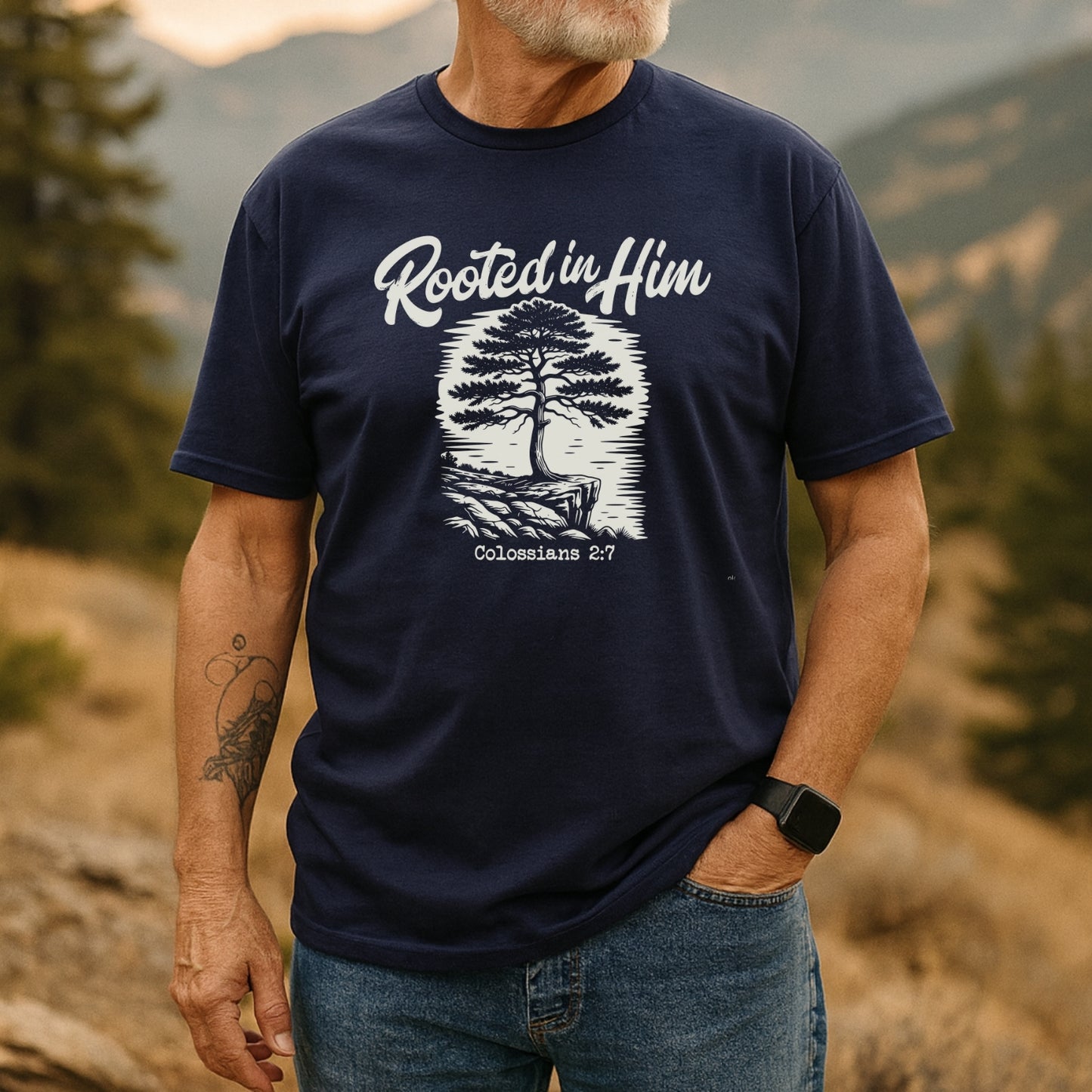 Man outdoors in a Rooted In Him Mountain T-Shirt, featuring a stylized tree and Colossians 2:7. Paired with jeans, he exudes adventure-ready comfort and timeless design.