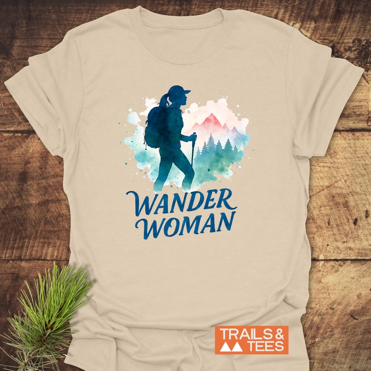 Wander Woman Watercolor T-Shirt featuring a graphic of a hiker with poles in front of trees and mountains on cream fabric, laid on wood with a pine sprig beside it.