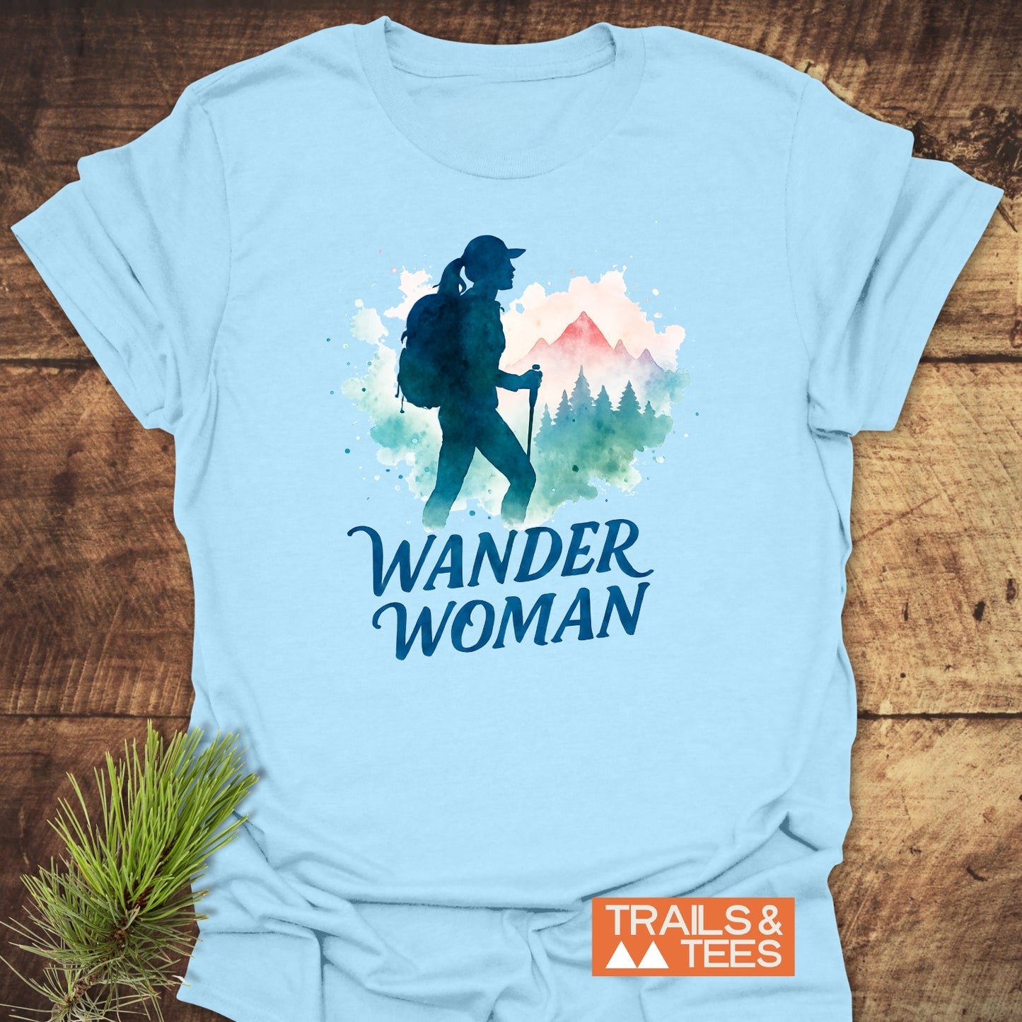 Wander Woman Watercolor T-Shirt featuring a hiker graphic with pine trees and mountains, laid on wood. Showcases a slogan and crafted from soft, durable cotton for outdoor enthusiasts.