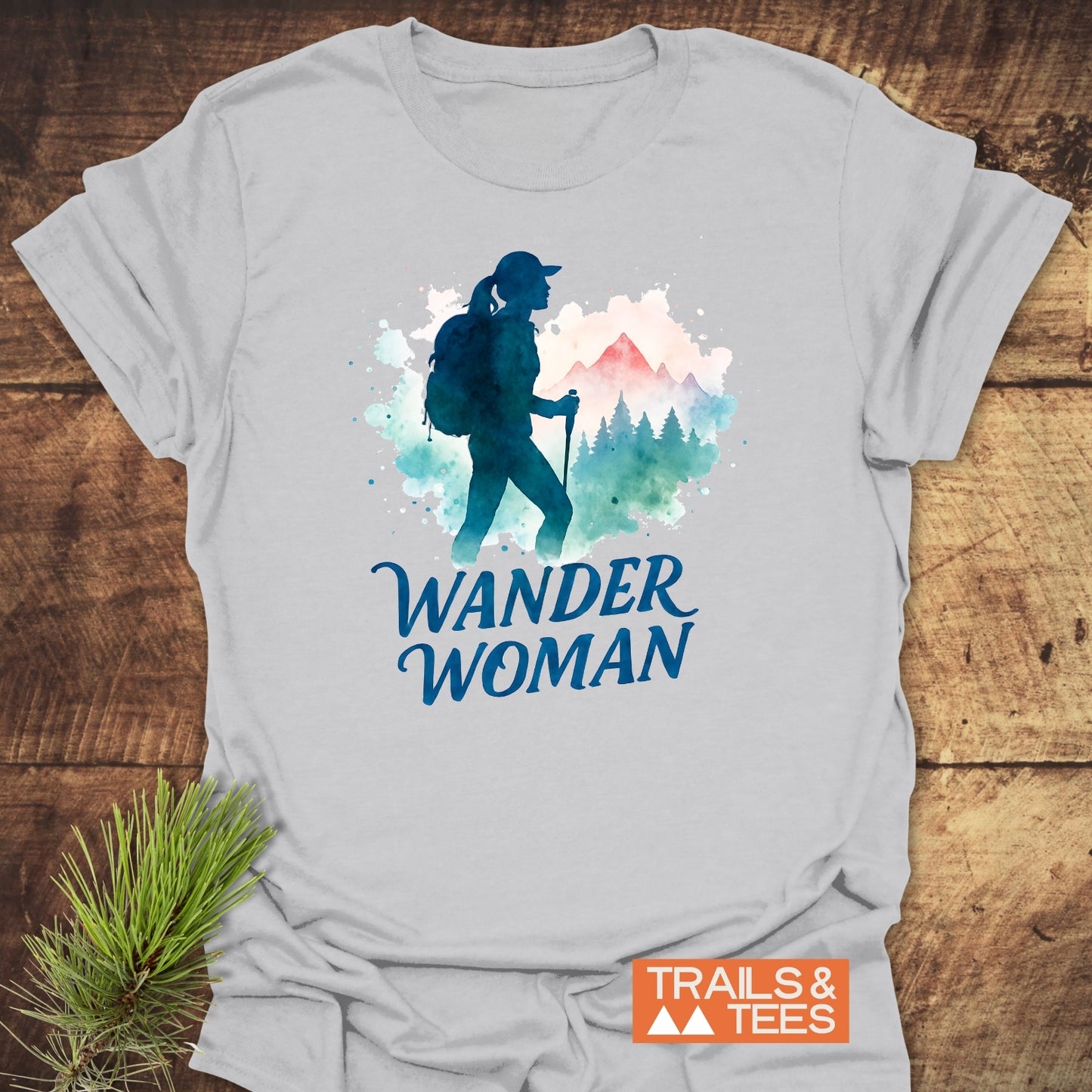 Wander Woman Watercolor T-Shirt featuring a silhouette of a woman hiker with a backpack and trekking pole against mountains and trees. Includes WANDER WOMAN text and TRAILS & TEES logo.