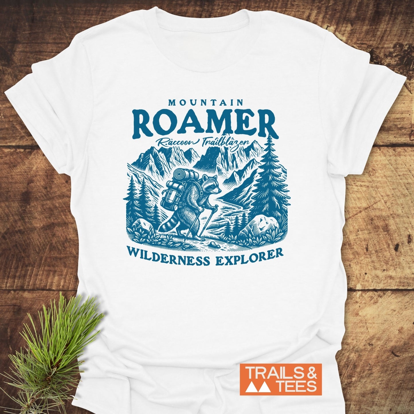 Mountain Roamer Raccoon T-Shirt featuring a blue illustration of a raccoon hiking through mountains, with captions MOUNTAIN ROAMER, Raccoon Trailblazer, and WILDERNESS EXPLORER, laid on a wooden surface.