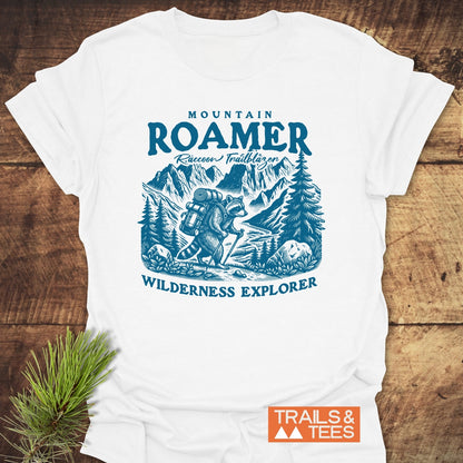 Mountain Roamer Raccoon T-Shirt featuring a blue illustration of a raccoon hiking through mountains, with captions MOUNTAIN ROAMER, Raccoon Trailblazer, and WILDERNESS EXPLORER, laid on a wooden surface.