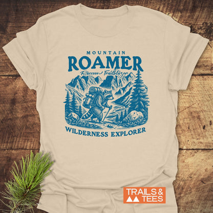 Mountain Roamer Raccoon T-Shirt featuring a blue hiking scene with a raccoon, backpack, and hiking stick. Text reads MOUNTAIN ROAMER,” Raccoon Trailblazer,” WILDERNESS EXPLORER. Trails & Tees logo included.