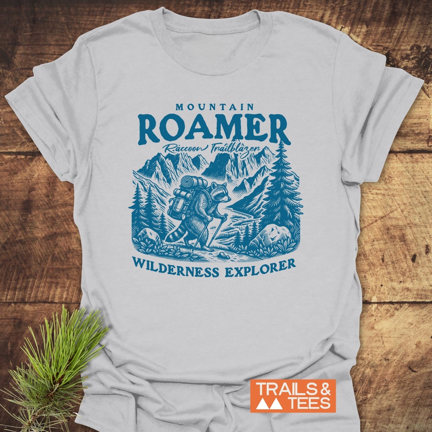 Mountain Roamer Raccoon T-Shirt featuring a blue illustration of a backpack-wearing raccoon hiking with trekking poles, set against a mountain stream backdrop with pine trees and peaks.