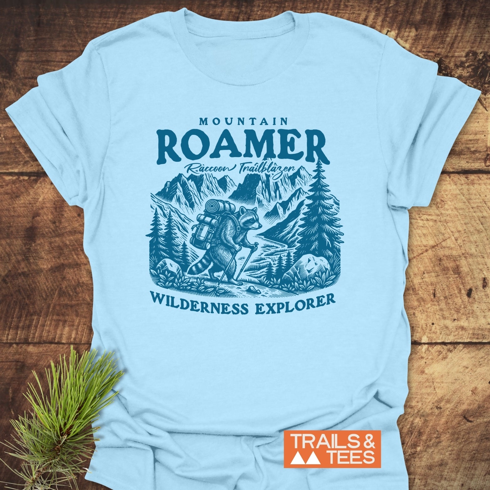 Mountain Roamer Raccoon T-Shirt features a teal raccoon illustration hiking with a backpack in a forest, surrounded by text Raccoon Trailblazer and Wilderness Explorer, showcasing adventure-ready design.