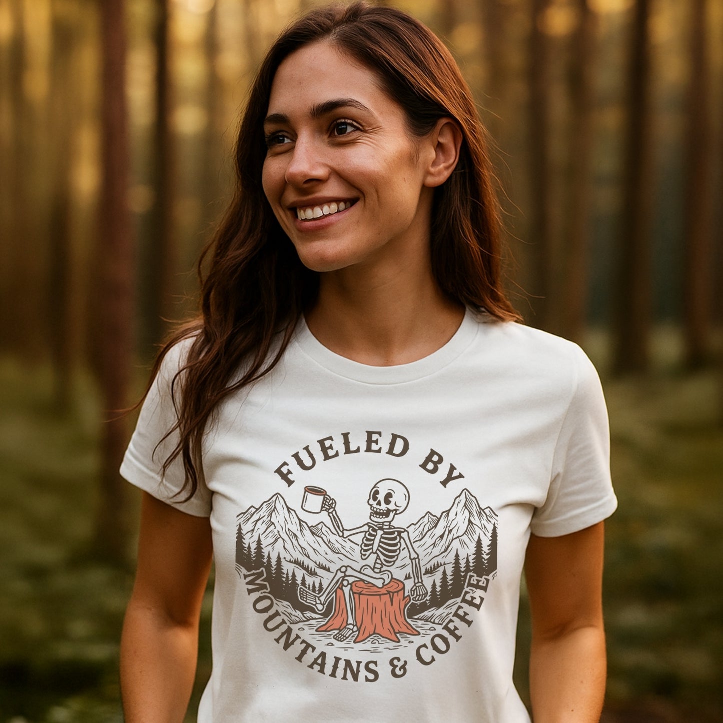 Woman in forest wearing Fueled By Mountains And Coffee T-Shirt featuring a skeleton with coffee on a stump, surrounded by mountains. Designed for comfort and adventure with durable prints and pre-shrunk fit.