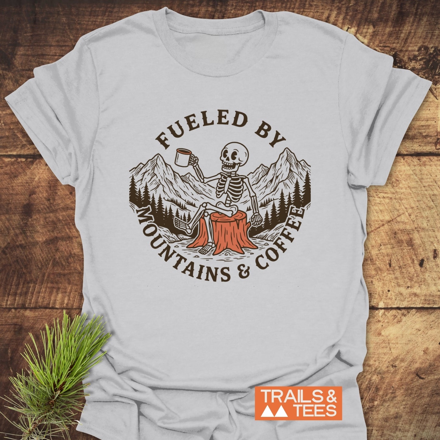 Fueled By Mountains And Coffee T-Shirt featuring a cartoon skeleton holding a coffee mug, sitting on a tree stump with mountain and tree graphics, laid on a wooden surface with a pine sprig.