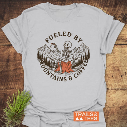 Fueled By Mountains And Coffee T-Shirt featuring a cartoon skeleton holding a coffee mug, sitting on a tree stump with mountain and tree graphics, laid on a wooden surface with a pine sprig.