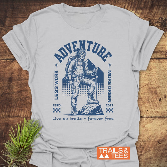 Adventure Retro T-Shirt featuring a backpacker graphic and motivational text, laid on wood. Made from soft cotton, it highlights outdoor enthusiasm with durable prints. Includes “TRAILS & TEES” label.