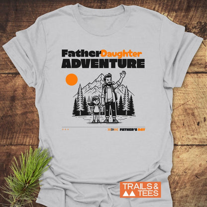 Father/Daughter Adventure T-Shirt featuring a cartoon of a dad and daughter by mountains and pines, with bold text and sun motif. Includes Father's Day call-out and TRAILS & TEES logo.