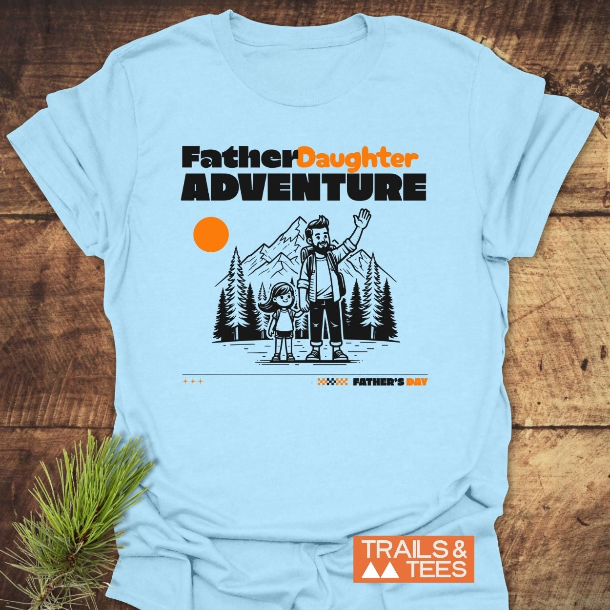 Father/Daughter Adventure T-Shirt features a cartoon of a father and daughter with pine trees and mountains, under the text “Father Daughter ADVENTURE.” Includes “Father’s Day” call-out and “TRAILS & TEES” logo.