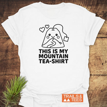 Mountain Tea Shirt T-Shirt on wooden surface, featuring a cartoon mountain-shaped tea bag graphic with hearts and “THIS IS MY MOUNTAIN TEA-SHIRT” slogan, embodying outdoor adventure and comfort.