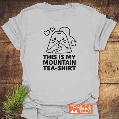 Mountain Tea Shirt T-Shirt laid on a wooden surface, featuring a smiling tea bag-mountain design with hearts and a slogan. Includes Trails & Tees logo. Crafted for comfort, ideal for outdoor enthusiasts.