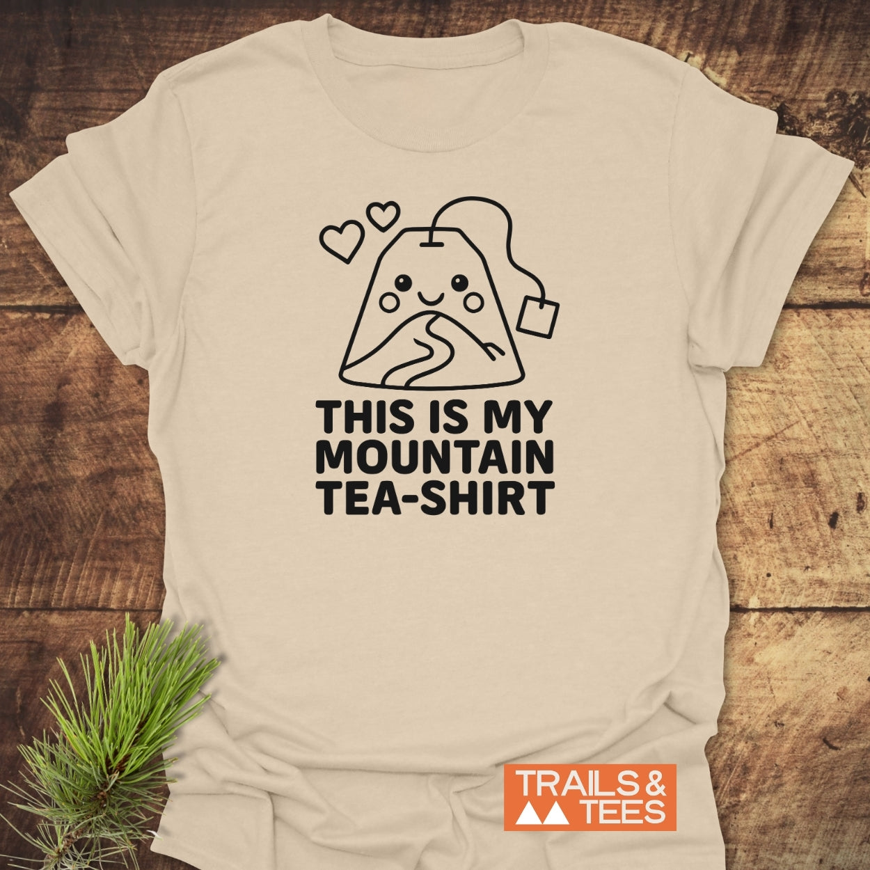 Mountain Tea Shirt T-Shirt features black line-art of a smiling, mountain-shaped tea bag with hearts, on soft beige cotton. Slogan reads “THIS IS MY MOUNTAIN TEA-SHIRT.” Orange “TRAILS & TEES” logo present.