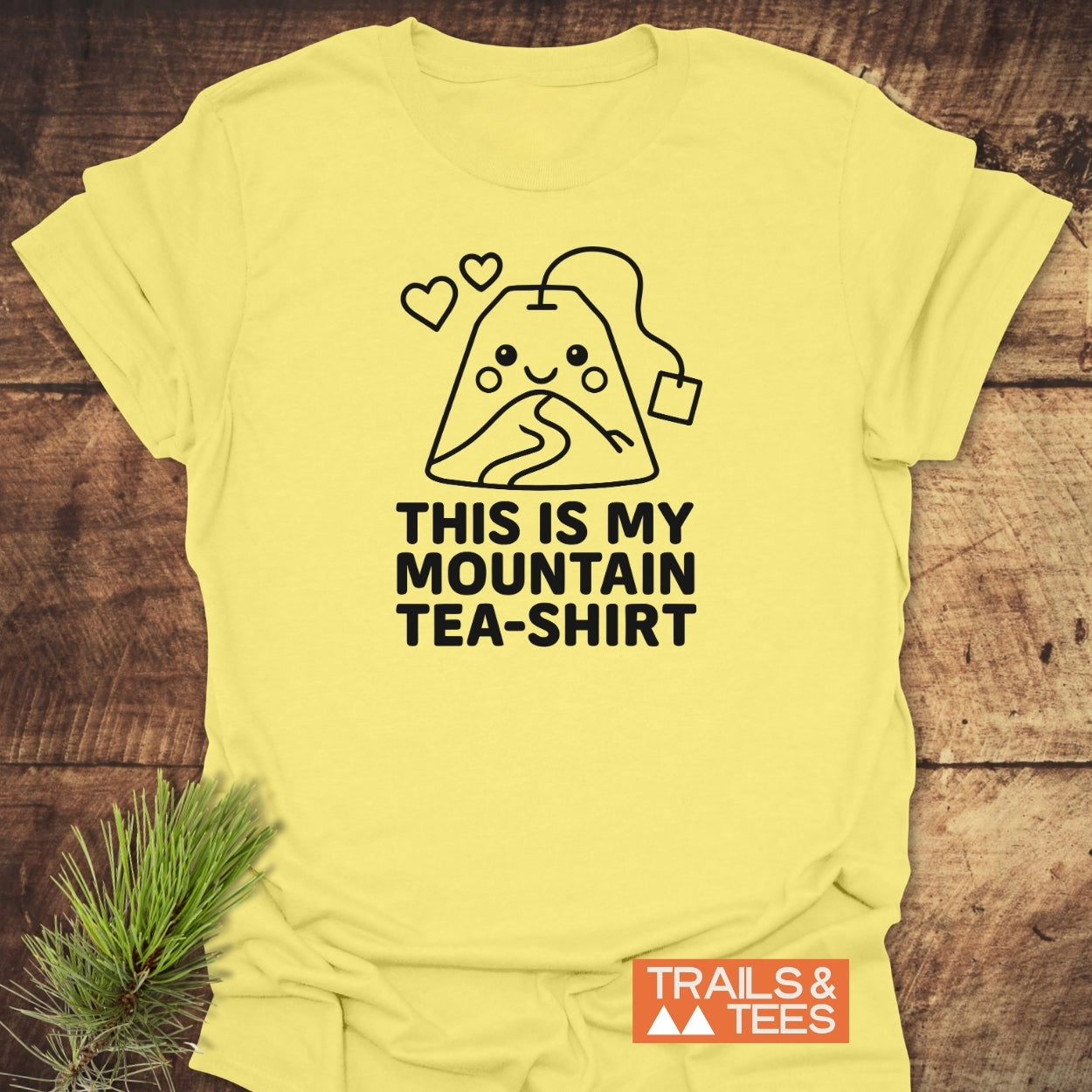 Mountain Tea Shirt T-Shirt features a black line-art design of a smiling tea bag like a mountain on a pale yellow tee, slogan “THIS IS MY MOUNTAIN TEA-SHIRT,” and a “TRAILS & TEES” logo.
