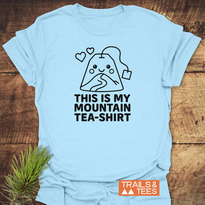 Mountain Tea Shirt T-Shirt featuring a black line drawing of a smiling tea bag with a mountain trail design and slogan “THIS IS MY MOUNTAIN TEA-SHIRT” on light-blue cotton, logo “TRAILS & TEES.”