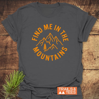 Find Me In The Mountains T-Shirt, featuring bold orange text and a mountain design, laid on wood with a pine sprig. Includes the TRAILS & TEES logo for an adventurous vibe.
