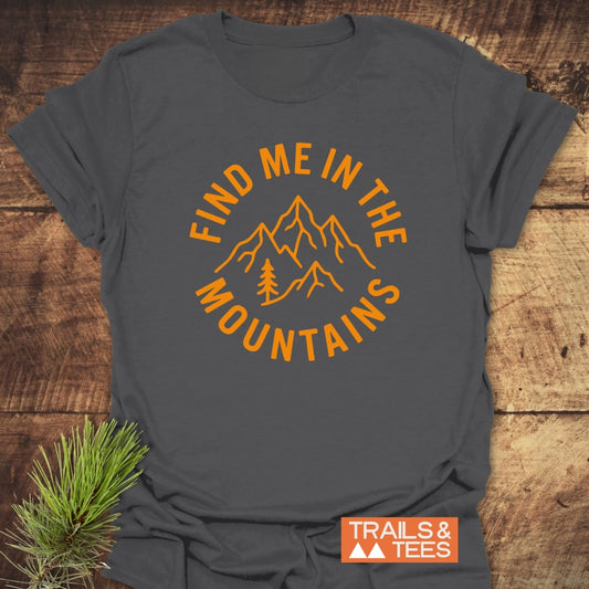 Find Me In The Mountains T-Shirt, featuring bold orange text and a mountain design, laid on wood with a pine sprig. Includes the TRAILS & TEES logo for an adventurous vibe.
